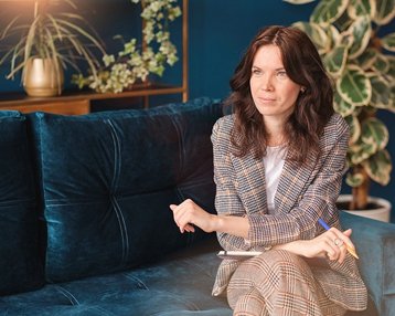 Eine Frau mit lockigem, braunem Haar sitzt auf einem blauen Sofa, trägt einen karierten Anzug und hält einen Stift. Sie schaut nachdenklich und ist in einem modernen Raum mit Pflanzen und dekorativen Elementen.